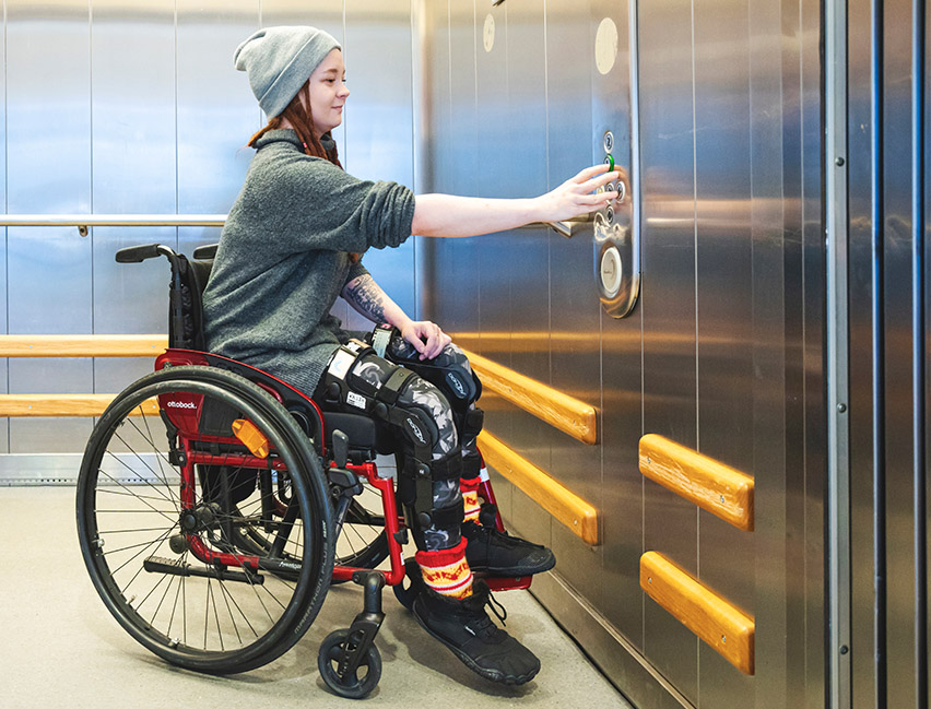 Nainen pyörätuolissa painamassa hissin kutsupainiketta.