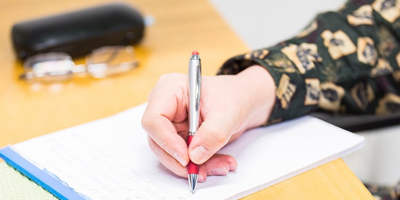 Nainen kirjoittaa kynällä ruutulehtiöön pöydän ääressä. Vieressä silmälasit ja -kotelo