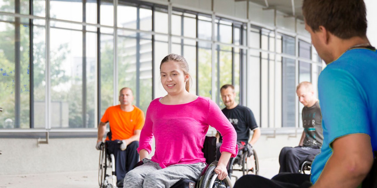 Nuori nainen harjoittelee pyörätuolilla liikkumista ja apuvälinekummi neuvoo vierestä.