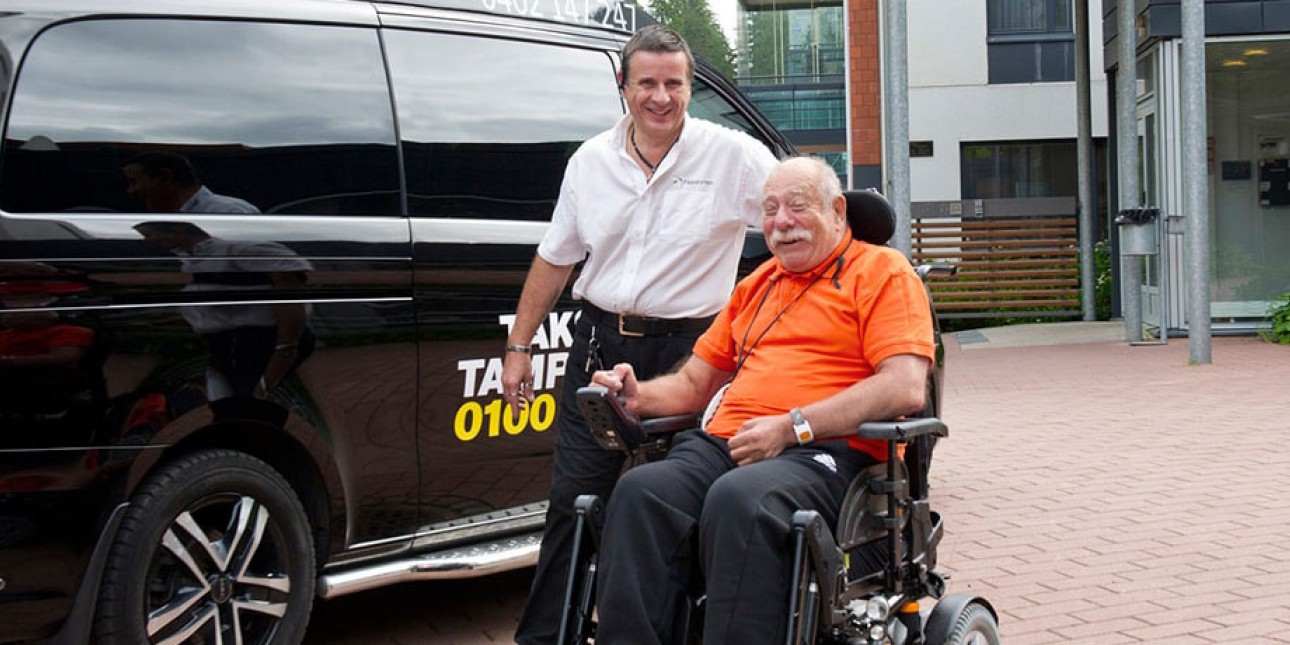 Mieskuljettaja auttaa pyörätuolissa istuvaa miesasiakasta taksiin.