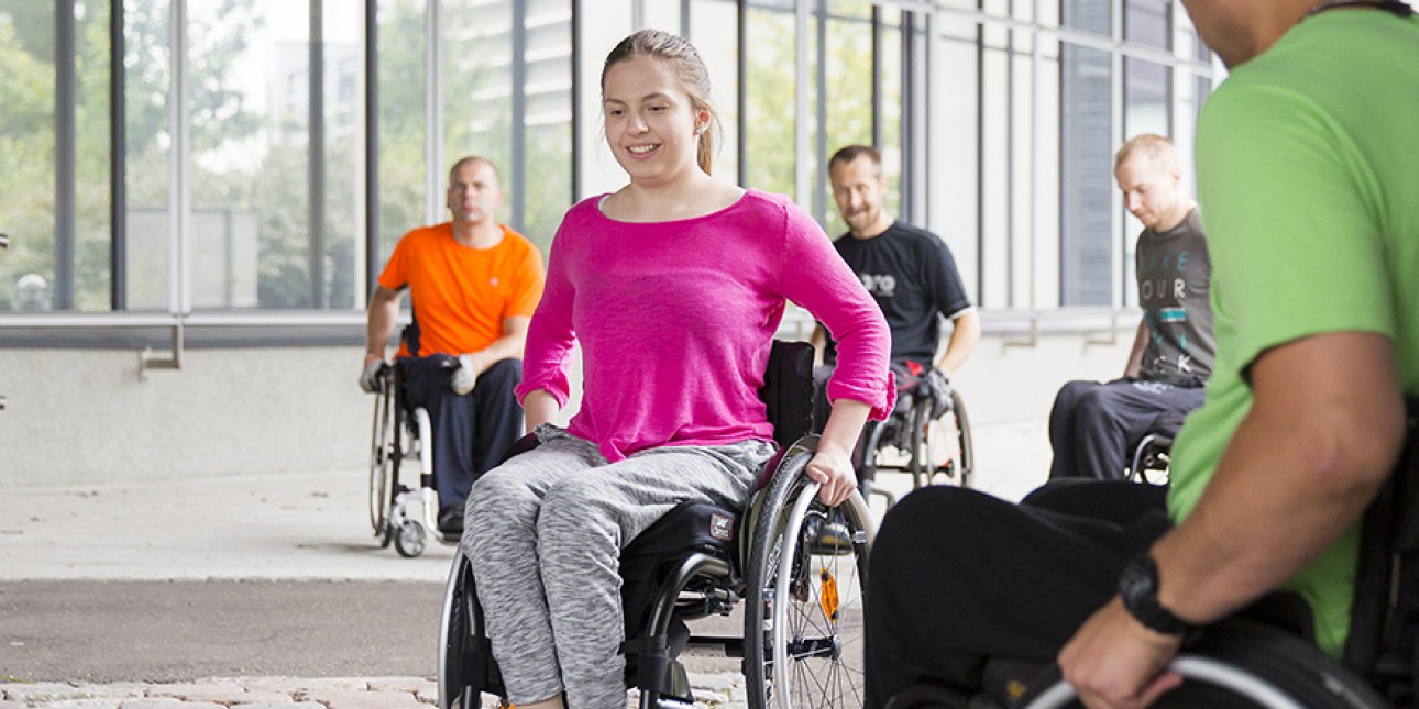 Pinkkipaitainen tyttö harjoittelee pyörätuolin käyttöä.