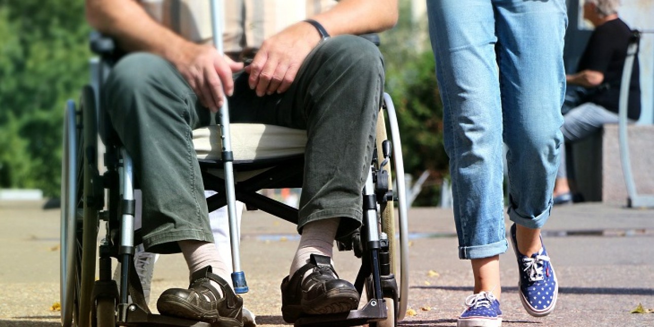 Alaosa pyörätuolissa istuvasta miehestä jolla kädessään kyynärsauva. Miehen vieressä alaosa kävelevästä farkkuhousuisesta naisesta lenkkareissa.