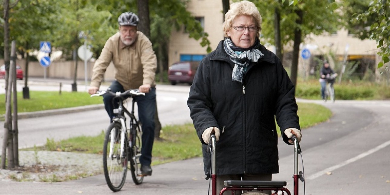 Vanhempi vaaleahiuksinen nainen mustassa takissa rollaattorin kanssa jalkakäytävän puolella yhdistetyllä pyörätiellä ja jalkakäytävällä. Pyörätien puolella ajaa takana vanhempi mies pyörällä pyöräilykypärä päässä. 
