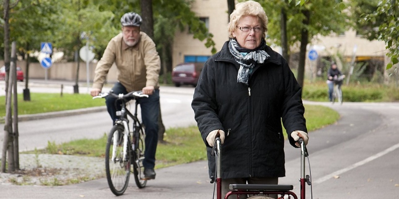 Vanhempi vaaleahiuksinen nainen mustassa takissa rollaattorin kanssa jalkakäytävän puolella yhdistetyllä pyörätiellä ja jalkakäytävällä. Pyörätien puolella ajaa takana vanhempi mies pyörällä pyöräilykypärä päässä.