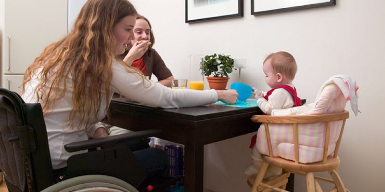 Ruokapöydässä nainen pyörätuolissa, vieressä lapsi syöttötuolissa, mies istuu naisen toisella puolella ja syö voileipää