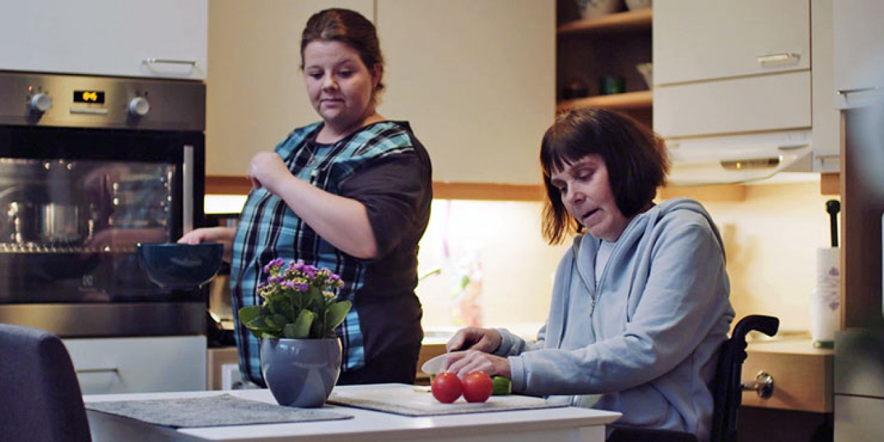 Keittiössä nainen pyörätuolissa leikkaa kurkkua, leikkuulaudalla myös 2 tomaattia. Hänen vieressään seisoo toinen nainen astia kädessä.