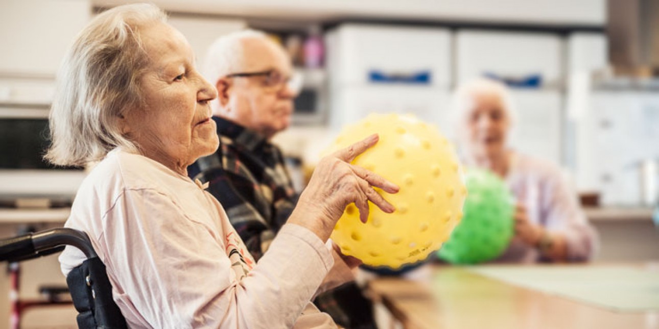 Etualalla vanha nainen pyörätuolissa pöydän äärellä keltainen jumppapallo kädessä, taustalla myös vanha mies ja nainen. Naisella vihreä pallo kädessä