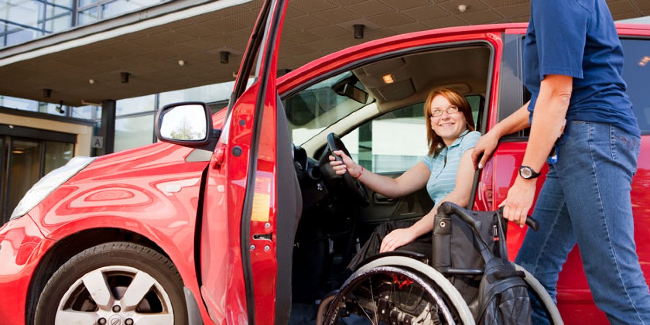 Punaisen henkilöauton kuljettajanpuoleinen ovi on auki ja penkillä istuu hymyilevä nainen. Mies tuö hänen viereensä pyörätuolia.