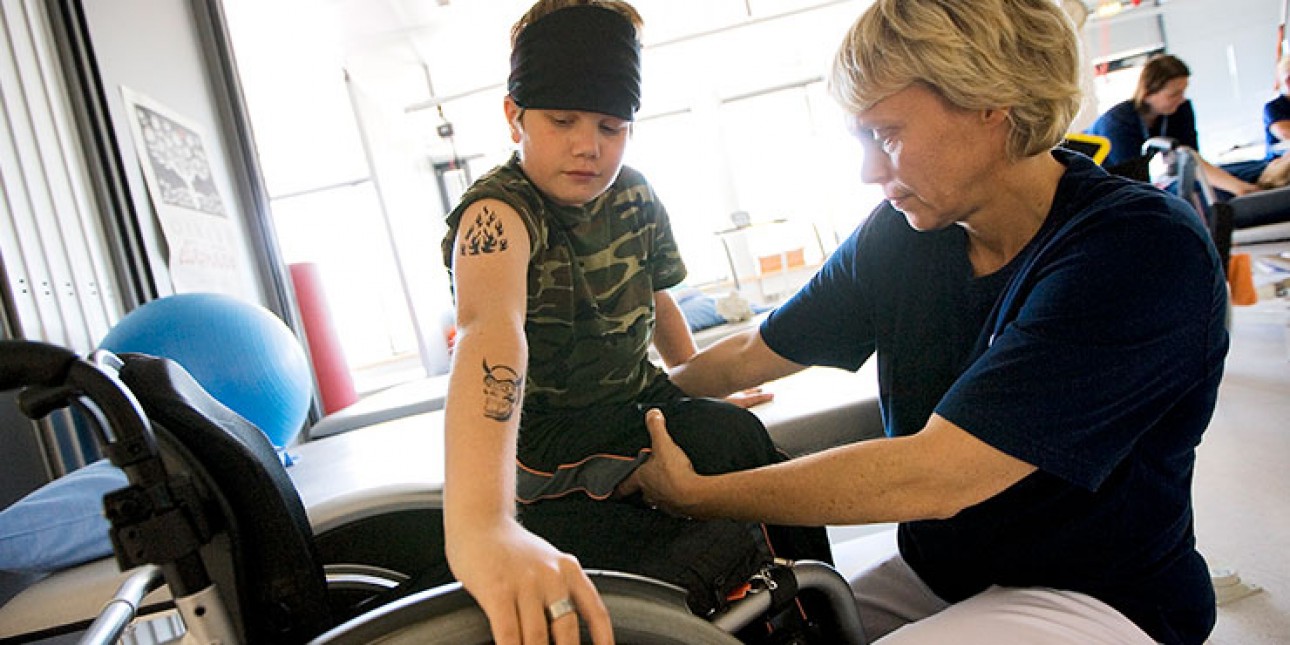 Terveydenhuollon ammattilainen auttaa poikaa istumaan pyörätuoliin. Kuvituskuva.