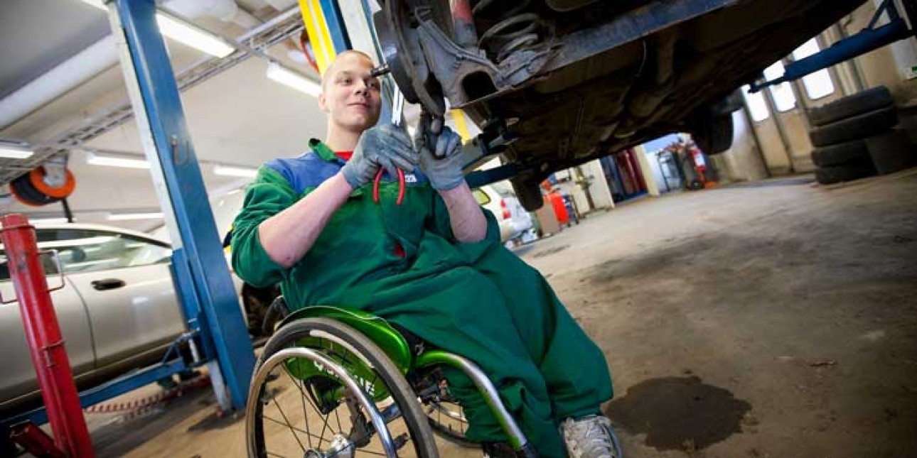 Pyörätuolissa istuva miesoletettu henkilö korjaa autoa. Auto on nostimen varassa. Korjaajalla on vihreä työhaalari ja harmaat hanskat.