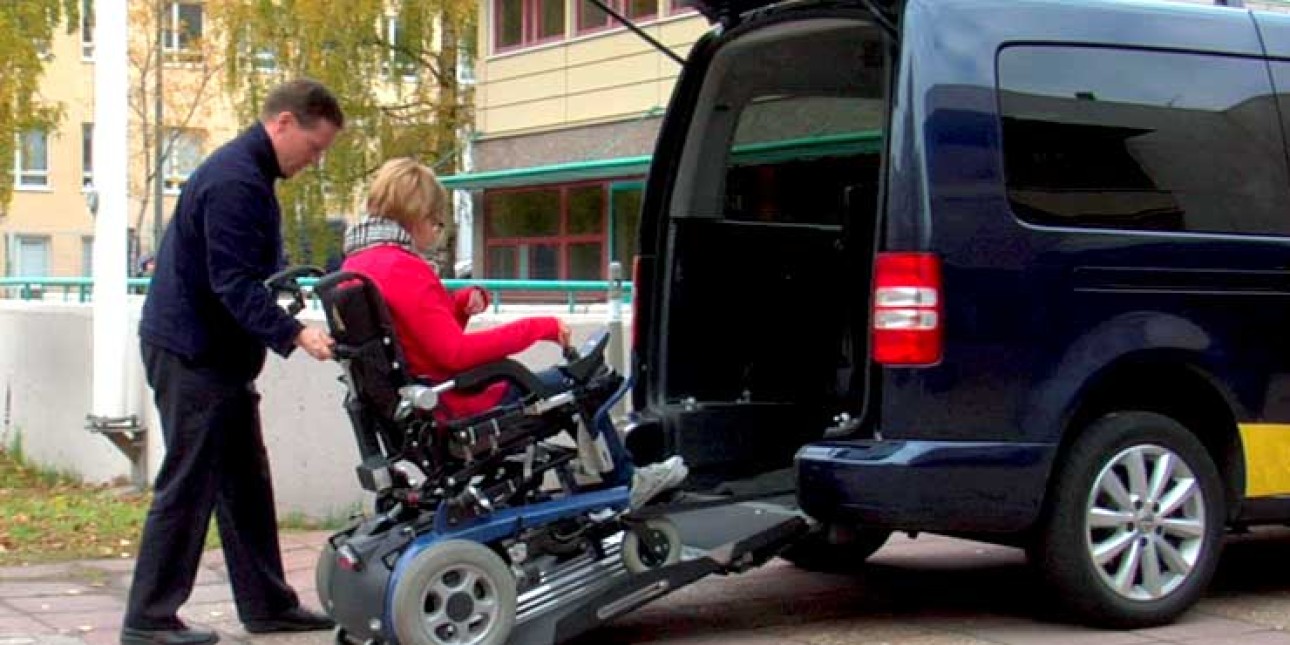 Taksinkuljettaja sirtää sähköpyörätuolia esteettömään taksiin.