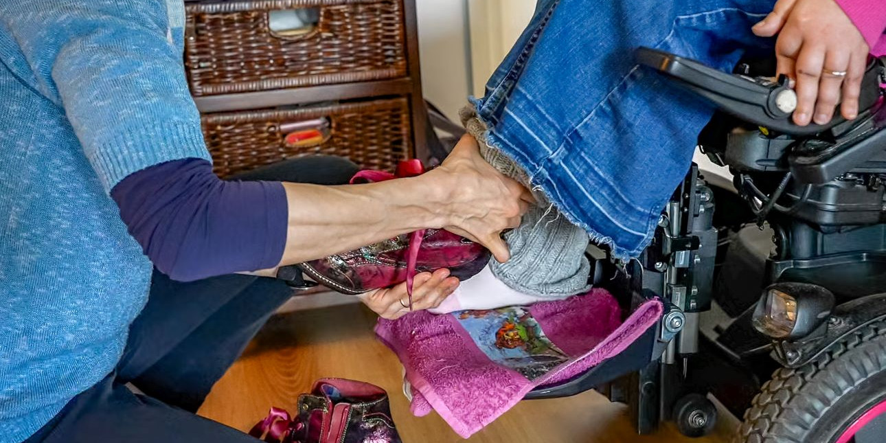 Avustaja auttaa avunkäyttäjälle kenkiä jalkaan