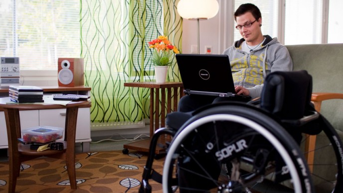 Nuori mies pyörätuolissa kotona tietokoneella. Kuvituskuva.