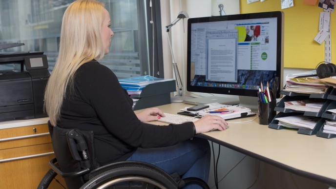 Vaaleahiuksinen nainen istuu pyörätuolissa tietokoneen ruudun äärellä