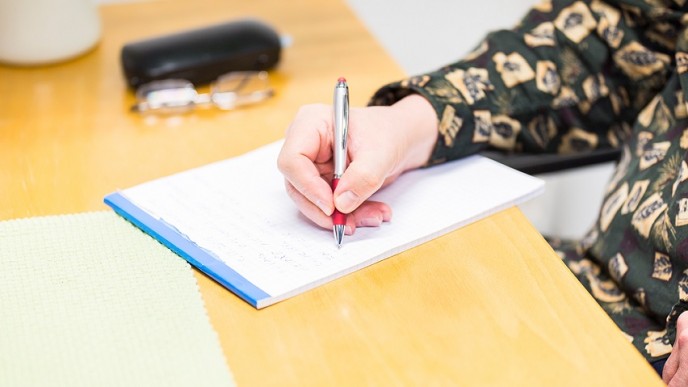 Nainen kirjoittaa kynällä ruutulehtiöön pöydän ääressä. Vieressä silmälasit ja -kotelo