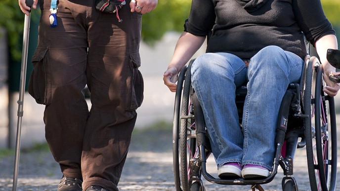Kuvassa mies kävelee kävelykeppiin tukeutuen. Miehen rinnalla nainen kelaa pyörätuolissa istuen ja avustajakoira kulkee vierellä.