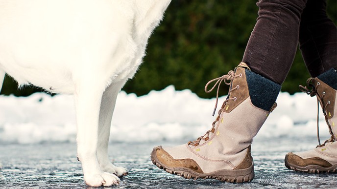 Liukkaalla tiellä seisomassa valkoinen labradorinnoutaja ja nainen. Näkyvissä vain koiran etutassut ja alaosaa sekä naisen kengät ja housut.
