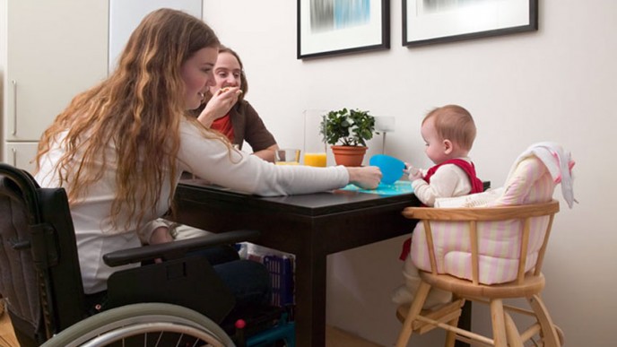 Ruokapöydässä nainen pyörätuolissa, vieressä lapsi syöttötuolissa, mies istuu naisen toisella puolella ja syö voileipää