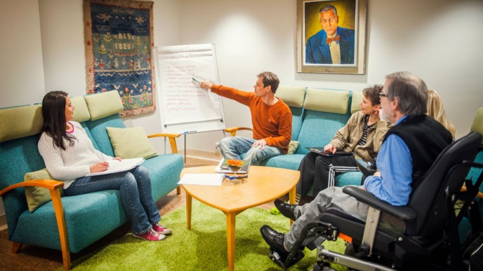 Neuvoja osoittaa fläppitaululle ja neuvottavat kuuntelevat nojatuoleissa ja apuvälineessä istuen.
