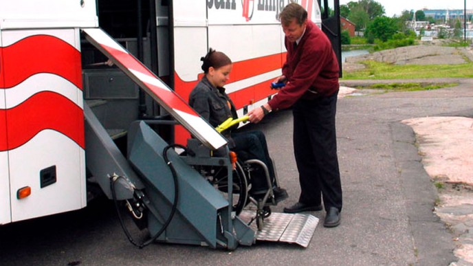 Kuljettaja auttaa pyörätuolissa istuvaa naista linja-autosta pois. Kuvituskuva.