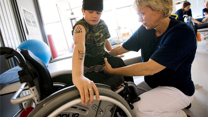 Terveydenhuollon ammattilainen auttaa poikaa istumaan pyörätuoliin. Kuvituskuva.