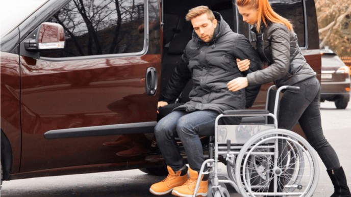 Henkilökohtainen avustaja auttaa pyörätuolissa istuvaa miestä autoon. Kuvituskuva.