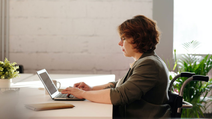 Pyörätuolissa istuva nainen työpöydän ääressä kirjoittamassa tietokoneella